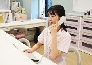 ピンク色のユニフォームを着た看護事務の画像です。受話器を耳に当て、入退院時の連絡をしているところです。